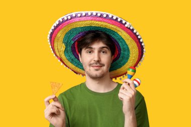 Sarı arka planda sombrero, marakas ve tortilla parçalı genç adam portresi. Ulusal Tortilla Cipsi Günü kutlaması