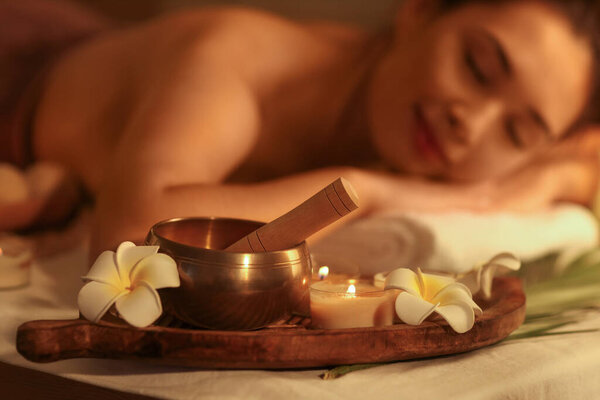 Тибетская чаша с цветами плюмерии и горящими свечами на диване в тёмном салоне спа, крупным планом