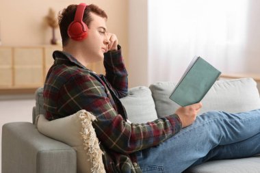 Genç adam oturma odasındaki kanepede müzik dinleyip kitap okuyor.