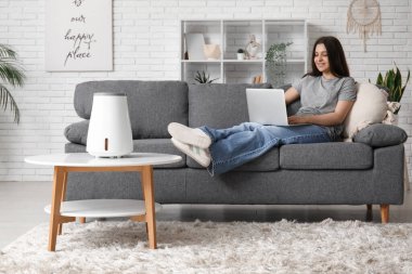 Genç bir kadın kanepede oturuyor ve modern nemlendiricinin yanında dizüstü bilgisayar kullanıyor oturma odasındaki sehpanın üzerinde.