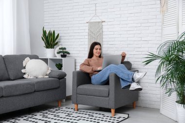 Genç bir kadın oturma odasındaki siyah koltukta dizüstü bilgisayar kullanıyor.