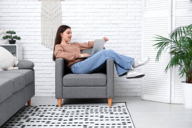 Genç bir kadın oturma odasındaki siyah koltukta dizüstü bilgisayar kullanıyor.