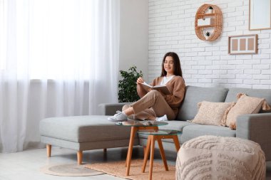 Genç kadın dergi okuyor ve oturma odasındaki gri kanepede dinleniyor.