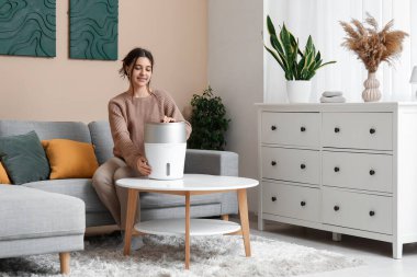 Genç bir kadın şık oturma odasında modern nemlendiriciyi açıyor.