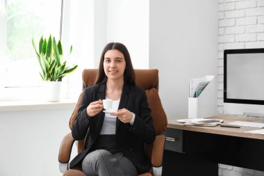 Bir fincan kahveyle çalışan bayan ofis çalışanı işyerindeki koltukta oturuyor.