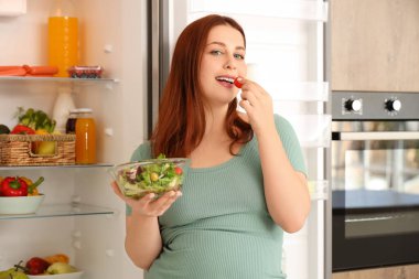 Genç hamile kadın mutfaktaki dolaptan taze sebze salatası alıyor.