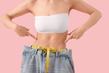 Pembe arka planda bol kot pantolon giyen genç ve güzel bir bayan, yakın plan. Kilo verme konsepti