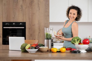 Not defteri ve blenderi olan sportif olgun bir kadın mutfakta smoothie yapıyor.