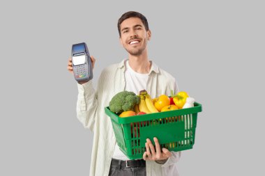 Gri arka planda alışveriş sepeti ve ödeme terminali olan genç bir adam.