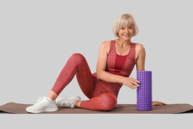 Gri arka planda yoga minderi ve köpük silindirli güzel, olgun, mutlu bir kadın.