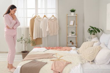 Düşünceli genç bir kadın yatak odasında kıyafet seçiyor.