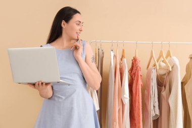 Bej arka planda dizüstü bilgisayarı olan düşünceli genç bir kadın.