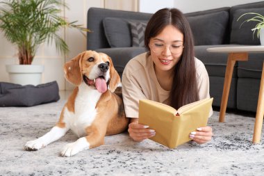 Gözlüklü genç kadın kitap okuyor ve oturma odasında yerde tapılası Beagle köpeğiyle yatıyor.