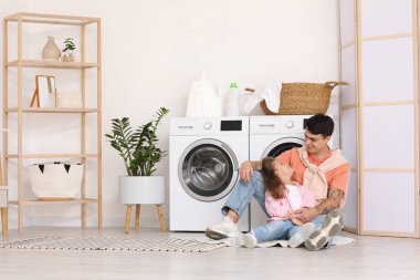 Genç baba ve kızı banyoda çamaşır makinesinin yanında oturuyorlar.