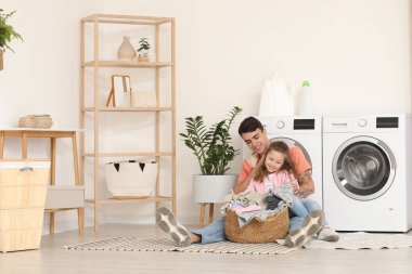 Genç baba ve kızı çamaşır sepetiyle banyoda çamaşır makinelerinin yanında oturuyorlar.