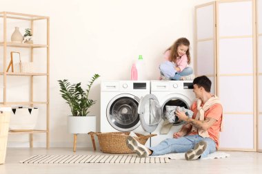 Genç baba ve kızı banyodaki çamaşır makinesine çamaşır koyuyorlar.