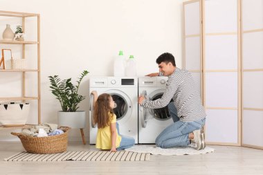 Genç baba ve kızı banyodaki çamaşır makinesine çamaşır koyuyorlar.