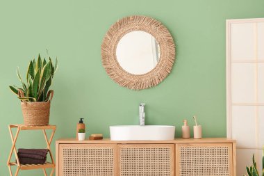 Seramik lavabo, ayna ve çekmeceler şık banyoda yeşil duvarın yanında.