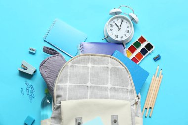 İçinde farklı kırtasiye malzemeleri, çalar saat ve mavi arka planda bir şişe su olan okul çantası.