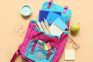 Farklı kırtasiye malzemeleriyle modern okul çantası, bej arka planda çalar saat ve elma.