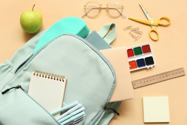 Farklı kırtasiye malzemeleri, gözlük ve bej arka planda elma ile modern okul sırt çantası.