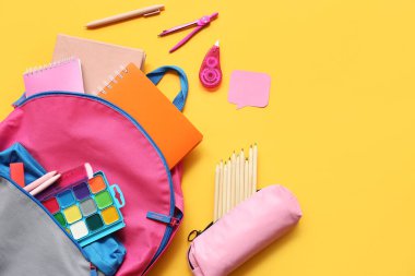 Sarı arka planda farklı kırtasiye malzemeleriyle modern okul sırt çantası.