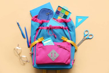 Bej arka planda farklı kırtasiye malzemeleri ve gözlüklerle modern okul sırt çantası.