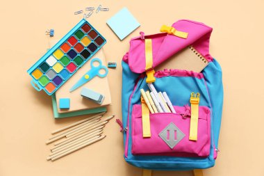 Bej arka planda farklı kırtasiye malzemesi içeren okul çantası.