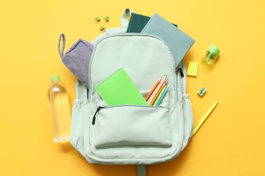 Okul çantasının içinde farklı kırtasiye malzemeleri ve sarı arka planda bir şişe su var.