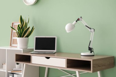 Ofiste yeşil duvarın yanında dizüstü bilgisayarı, saksısı ve lambası olan bir işyeri.