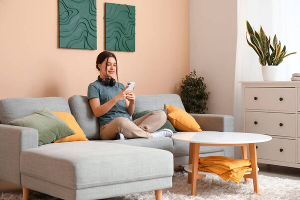 Young woman in headphones chatting with mobile phone and resting on comfortable sofa in living room