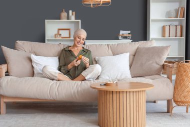 Kemoterapi üstüne kemoterapi ve bir şişe otla evde koltukta oturan olgun bir kadın.