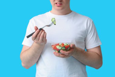 Mavi arka planda bir kase sağlıklı salatası olan şişman genç adam, yakın plan. Kilo alma kavramı