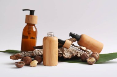 Beyaz arka planda Macadamia fındıkları, yeşil yaprak, ağaç kabuğu ve kozmetik ürünler şişeleri