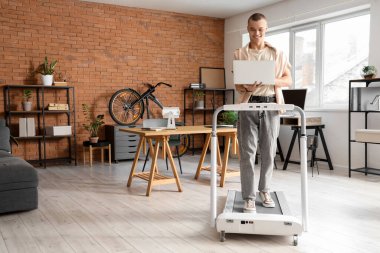 Genç işadamı ofisteki koşu bandında dizüstü bilgisayarla çalışıyor.