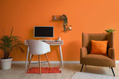 Modern bilgisayar ve turuncu duvarın yanındaki koltuk ile rahat çalışma alanı