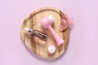 Ahşap tepsi, yüz masajı aletleri, kavanoz ve pembe arka planda bir şişe kozmetik ürünü.