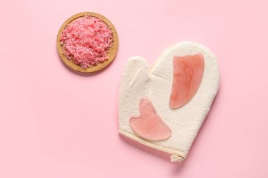 Yüz masajı aletleri, masaj eldiveni ve pembe arka planda bir kase deniz tuzu.