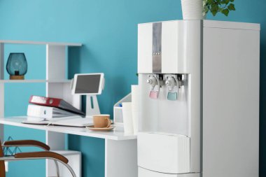 Ofisteki mavi duvarın yanındaki masada modern su soğutucusu ve kağıt bardak.