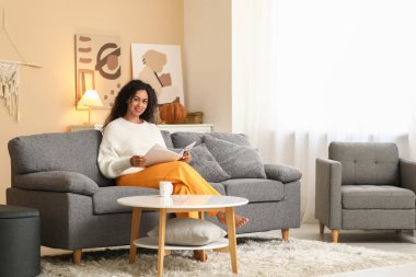 Evdeki kanepede çay okuyan güzel, mutlu, Afro-Amerikan kadın.