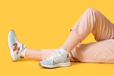 Spor ayakkabılı kadın bacakları, sarı arka planda yerde yatıyor.