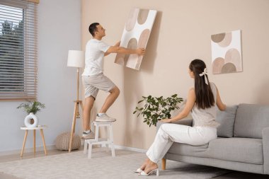 Genç bir kadın kanepede oturuyor ve kocası evdeki bej duvarlara resim asıyor.