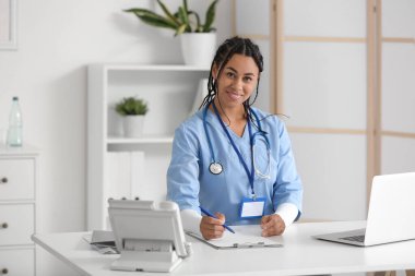 Laptop ve not panosuyla klinikteki masada oturan genç Afro-Amerikalı kadın doktor.