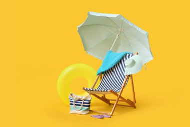 Güverte sandalyesi, plaj şemsiyesi, çanta, şapka, havlu, parmak arası terlik ve sarı arka planda şişme yüzük.