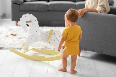 Küçük kız, annesiyle birlikte yürümeyi öğreniyor. Sallanan atın yanında, arka planda.