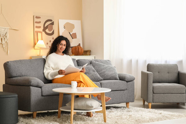 Beautiful young happy African-American woman in autumn outfit with cup of tea reading magazine on sofa at home