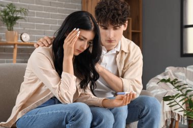 Evdeki kanepede oturan, hamilelik testi olan üzgün genç bir çift.