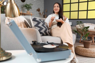 Evdeki kanepede oturan kahve fincanı ve pikabı olan genç bir kadın.