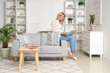 Evdeki kanepede dizüstü bilgisayar kullanan olgun bir kadın.