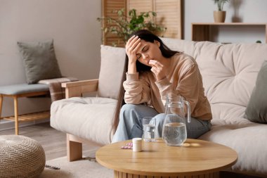 Kanepede oturan ve evinde su ve haplarla baş ağrısı çeken genç bir kadın.